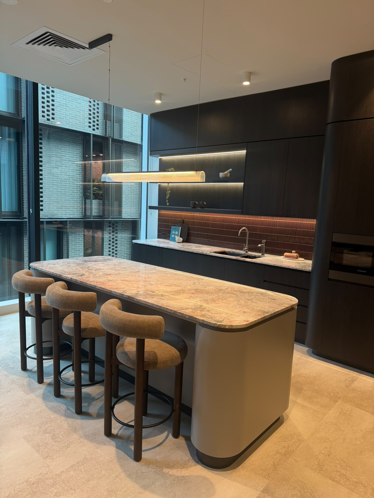 Rounded stone island with upholstered stools, dark cabinets and warm pendant lighting